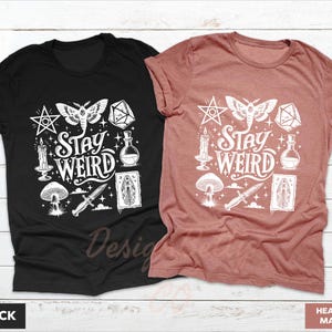 May include: Black and heather mauve t-shirts with a white graphic design. The design includes a moth, pentagram, dice, candle, mushroom, potion bottle, dagger, and the words "Stay Weird". The shirts are displayed on a white wooden surface.