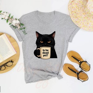 K&ouml;nnte beinhalten: Hellgraues T-Shirt mit einer schwarzen Katze, die eine Kaffeetasse mit der Aufschrift "Is he dead yet?" h&auml;lt.