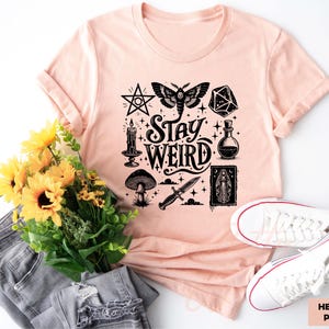 May include: Heather peach t-shirt with a black graphic that says "Stay Weird" and features a pentagram, moth, dice, potion bottle, candle, mushroom, dagger, and a religious icon. The shirt is paired with jeans and white sneakers.