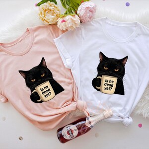 K&ouml;nnte beinhalten: Zwei T-Shirts mit einer schwarzen Katze, die eine Kaffeetasse mit der Aufschrift "Is he dead yet?" h&auml;lt. Ein Shirt ist in der Farbe Heather Peach und das andere ist wei&szlig;.
