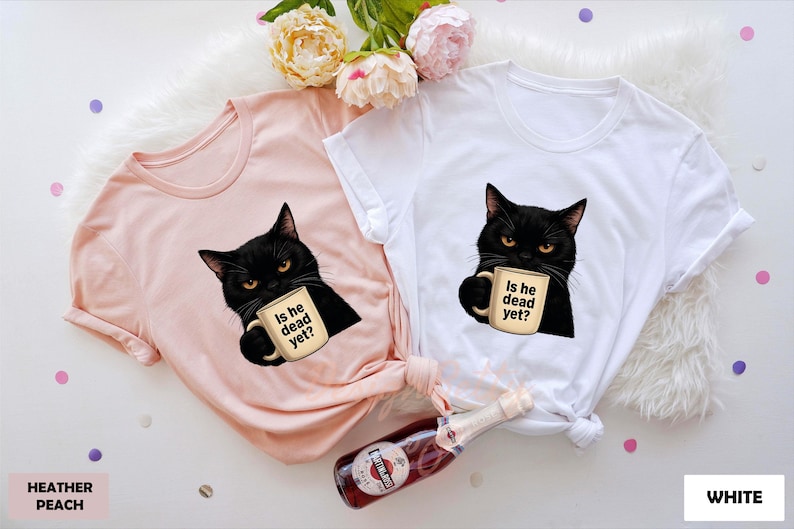 K&ouml;nnte beinhalten: Zwei T-Shirts mit einer schwarzen Katze, die eine Kaffeetasse mit der Aufschrift "Is he dead yet?" h&auml;lt. Ein Shirt ist in der Farbe Heather Peach und das andere ist wei&szlig;.