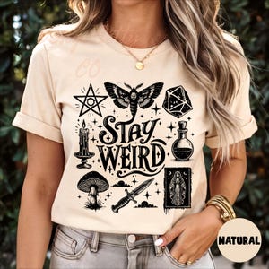 May include: Beige t-shirt with a black graphic design featuring a moth, pentagram, mushroom, dagger, and the words "Stay Weird." The design also includes a candle, potion bottle, and a die. The shirt is made of a soft material.