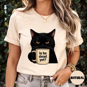 K&ouml;nnte beinhalten: Eine Frau tr&auml;gt ein beiges T-Shirt mit einer schwarzen Katze, die eine Kaffeetasse mit der Aufschrift "Is he dead yet?" h&auml;lt. Die Katze hat einen m&uuml;rrischen Gesichtsausdruck.