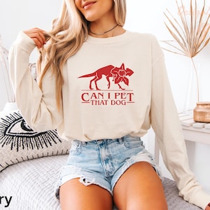A woman wearing a white long-sleeved shirt with a red graphic and text, sitting on a bed with a black and white pillow.