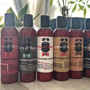 May include: Six bottles of beard conditioner with black caps and maroon liquid. Each bottle has a label with a bearded man wearing sunglasses and a bow tie. The text on the label reads "Bosses & Bean Grooming Lounge".