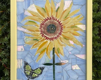 May include: A mosaic art piece featuring a large yellow sunflower with a brown center and a green butterfly. The artwork is set against a light blue background and framed in a yellow rectangular frame.