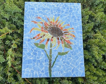 May include: A mosaic artwork depicting a sunflower. The flower has a brown center, yellow, orange, and brown petals, and green leaves and stem. The background is light blue, made of small mosaic tiles.