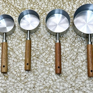 May include: A set of four stainless steel measuring cups with wooden handles. Each cup has a different capacity, with measurements in cups and milliliters printed on the handles. The cups are arranged in a row.