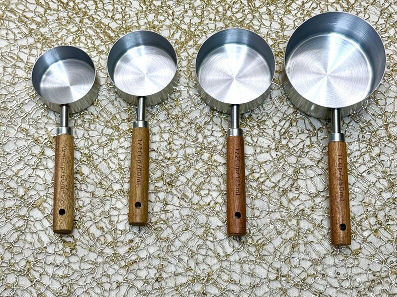May include: A set of four stainless steel measuring cups with wooden handles. Each cup has a different capacity, with measurements in cups and milliliters printed on the handles. The cups are arranged in a row.