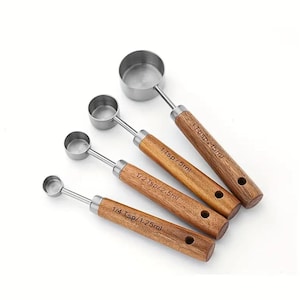 May include: A set of four stainless steel measuring spoons with wooden handles. Each spoon is labeled with its measurement in teaspoons and milliliters: 1 Tbsp/15ml, 1 Tsp/5ml, 1/2 Tsp/2.5ml, and 1/4 Tsp/1.25ml. The handles have a natural wood grain.