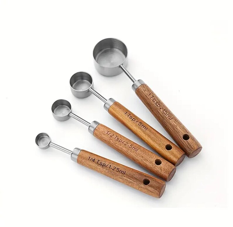 May include: A set of four stainless steel measuring spoons with wooden handles. Each spoon is labeled with its measurement in teaspoons and milliliters: 1 Tbsp/15ml, 1 Tsp/5ml, 1/2 Tsp/2.5ml, and 1/4 Tsp/1.25ml. The handles have a natural wood grain.