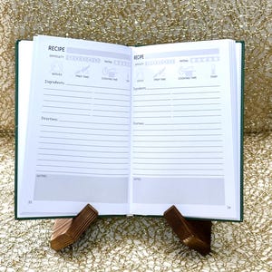 May include: An open recipe journal with green covers, displayed on a wooden stand. The pages are white with printed sections for ingredients, directions, and notes. The text "RECIPE" is visible at the top of each page.