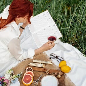 May include: A person is reading a recipe book while enjoying a cup of tea during a picnic. A wooden tray holds lemons, grapefruit slices, cinnamon sticks, walnuts, and a small plate. The scene is set on a white blanket with a backdrop of green grass.