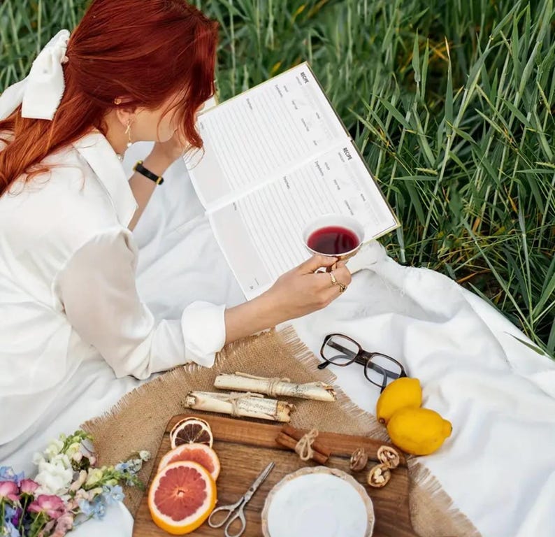 May include: A person is reading a recipe book while enjoying a cup of tea during a picnic. A wooden tray holds lemons, grapefruit slices, cinnamon sticks, walnuts, and a small plate. The scene is set on a white blanket with a backdrop of green grass.