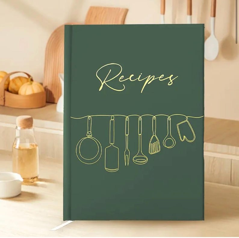 May include: A dark green recipe book with the word "Recipes" in gold script. Below the word, a gold line drawing of kitchen utensils hangs across the cover. A white ribbon bookmark is visible.