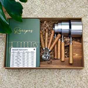 May include: A boxed set of kitchen tools includes a green recipe book, measuring cups, and measuring spoons. The measuring cups are stainless steel, and the spoons have wooden handles. A measurement conversion chart is included.