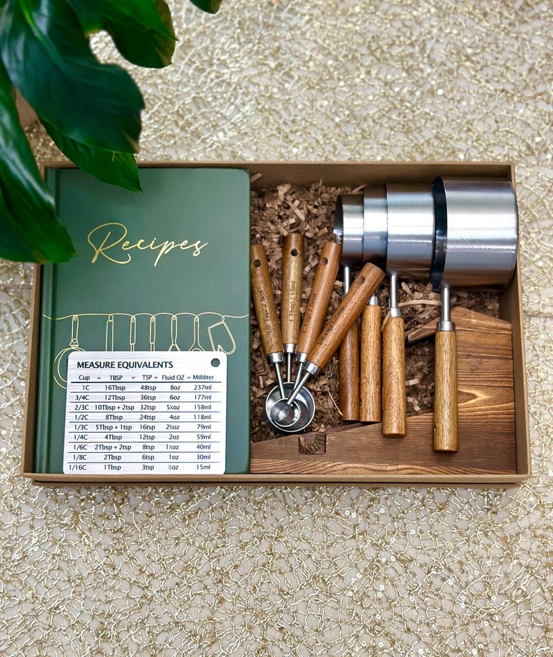 May include: A boxed set of kitchen tools includes a green recipe book, measuring cups, and measuring spoons. The measuring cups are stainless steel, and the spoons have wooden handles. A measurement conversion chart is included.