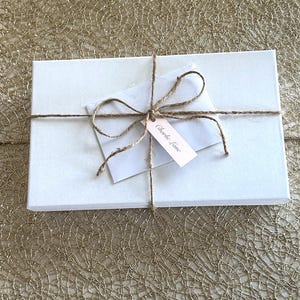 May include: A white rectangular gift box tied with twine. A small white envelope and a pink tag with the text "Charlie Lane" are attached to the twine. The box is on a gold textured surface.