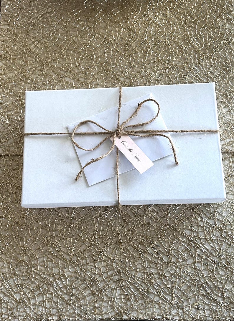 May include: A white rectangular gift box tied with twine. A small white envelope and a pink tag with the text "Charlie Lane" are attached to the twine. The box is on a gold textured surface.
