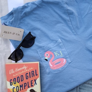 May include: A light blue t-shirt with a pocket featuring a pink flamingo pool float with the initials "KJ" embroidered on it. The t-shirt is lying on a white and gray striped towel next to a book titled "Good Girl Complex" by Elle Kennedy and a pair of black sunglasses.