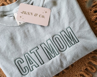 Puede incluir: Una camiseta azul claro con el texto "CAT MOM" bordado en verde. La camiseta está doblada y sobre una estera tejida marrón. Una etiqueta rosa con el texto "JKSN & CO." está unida a la camiseta.