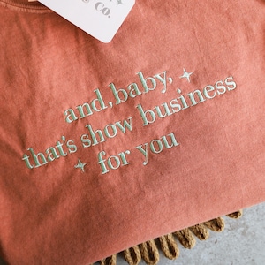 Puede incluir: Una camiseta de color coral con la frase bordada "and, baby, that's show business for you" en verde claro. La camiseta tiene cuello redondo y una etiqueta que dice "JKSN & CO." La camiseta est&aacute; doblada sobre una superficie tejida.