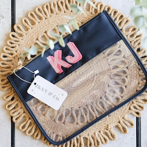 May include: A black and clear zippered pouch with a pink and white embroidered "KJ" on the front. The pouch is lying on a woven straw mat. A white tag with the text "JKSN & Co." is attached to the pouch.