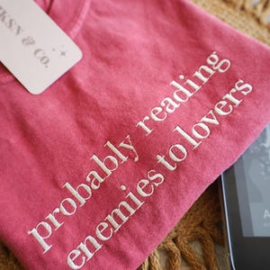 May include: A pink t-shirt with the text "probably reading enemies to lovers" embroidered in white.