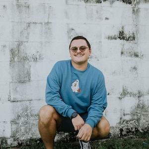 May include: A man wearing a blue sweatshirt with a ghost graphic and a pair of black-framed glasses is squatting in front of a white brick wall. He is wearing black shorts and black sneakers.
