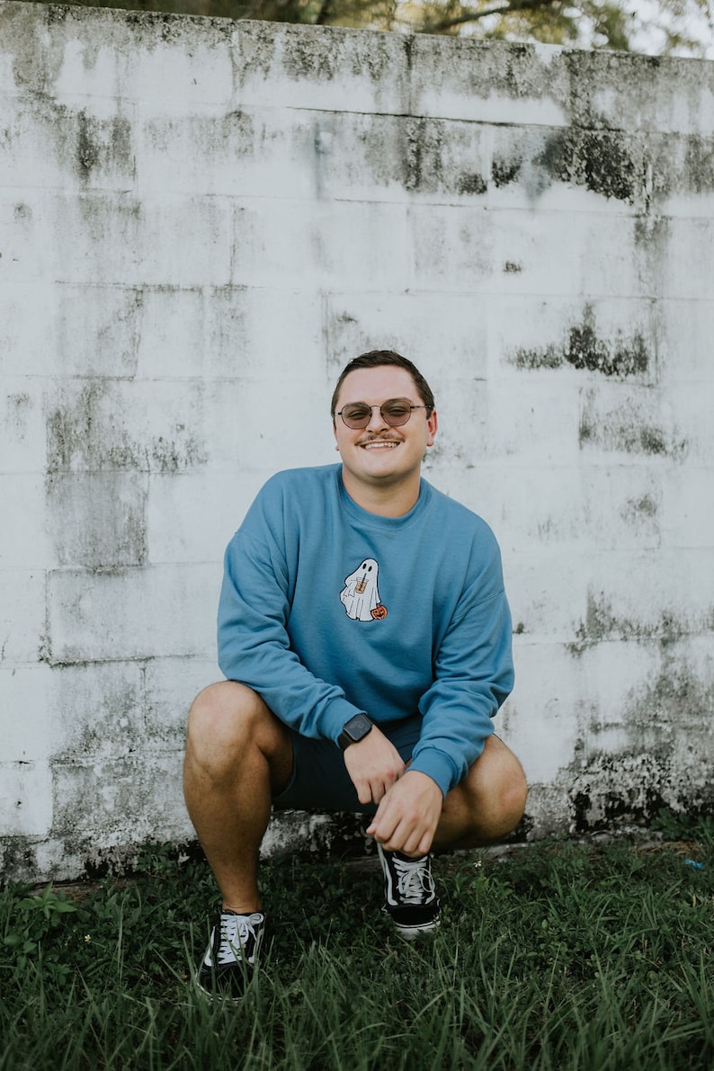 May include: A man wearing a blue sweatshirt with a ghost graphic and a pair of black-framed glasses is squatting in front of a white brick wall. He is wearing black shorts and black sneakers.