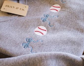 May include: Gray sweatshirt with a row of embroidered baseballs and blue bows. The sweatshirt has a tag that says "JKSN & CO."