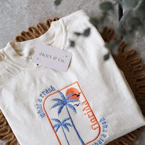 May include: A white t-shirt with an embroidered design of palm trees and a sunset. The text "Florida" is written vertically on the right side of the design. The text "what a crash what a rush" is written on the left side of the design. The t-shirt has a tag that says "JKSN & Co."