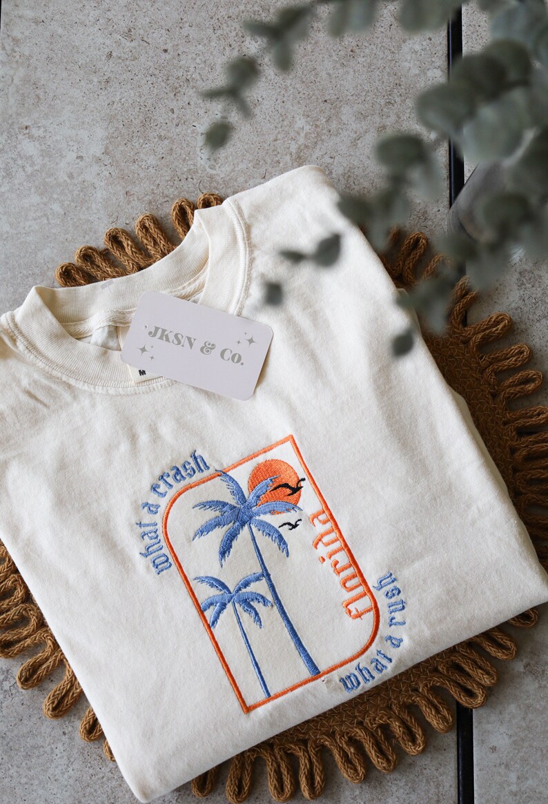May include: A white t-shirt with an embroidered design of palm trees and a sunset. The text "Florida" is written vertically on the right side of the design. The text "what a crash what a rush" is written on the left side of the design. The t-shirt has a tag that says "JKSN & Co."