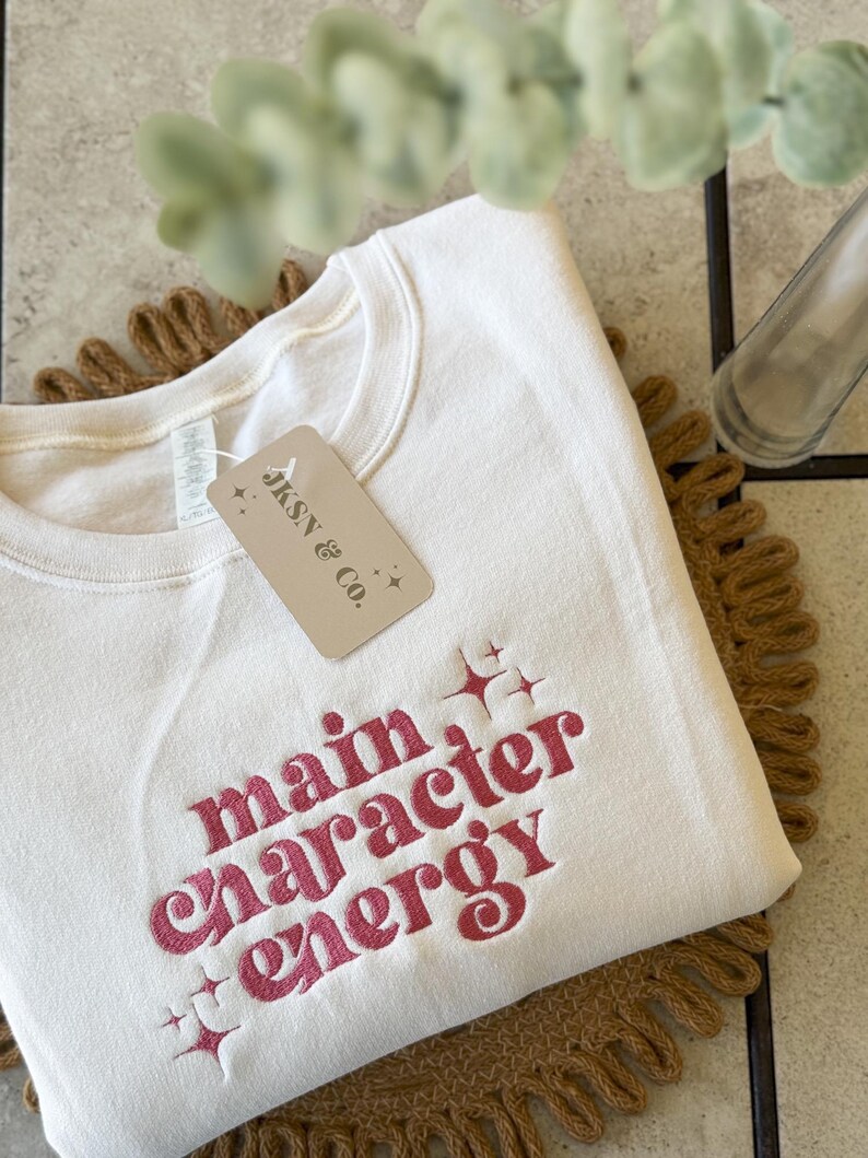 May include: A cream colored sweatshirt with the text "main character energy" embroidered in pink. The text is in a retro font with stars around it. The sweatshirt has a tag that says "JKSN & Co."