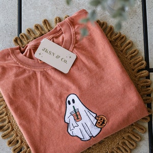 Może przedstawiać: Pomarańczowy t-shirt z haftowanym duchem trzymającym latte z przyprawą dyniową i małą dynią. Na koszulce znajduje się metka z napisem "JKSN & CO."