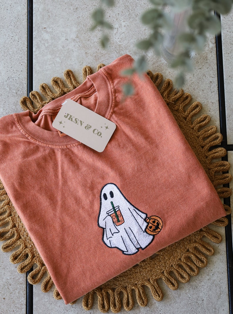 Może przedstawiać: Pomarańczowy t-shirt z haftowanym duchem trzymającym latte z przyprawą dyniową i małą dynią. Na koszulce znajduje się metka z napisem "JKSN & CO."