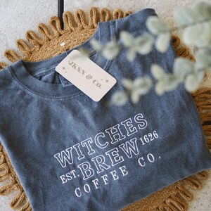 Puede incluir: Una camiseta gris oscuro con el texto "Witches Brew Est. 1626 Coffee Co." en letras blancas. La camiseta est&aacute; doblada y sobre una estera tejida marr&oacute;n.