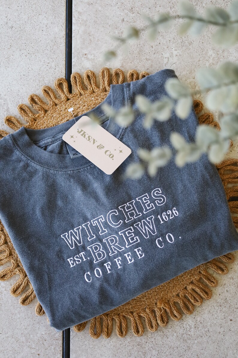 Puede incluir: Una camiseta gris oscuro con el texto "Witches Brew Est. 1626 Coffee Co." en letras blancas. La camiseta est&aacute; doblada y sobre una estera tejida marr&oacute;n.