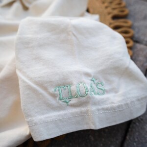 Puede incluir: Primer plano de una manga de camiseta color crema. El texto "TLOAS" est&aacute; bordado en color verde claro en la manga. La camiseta est&aacute; doblada y el fondo est&aacute; borroso.