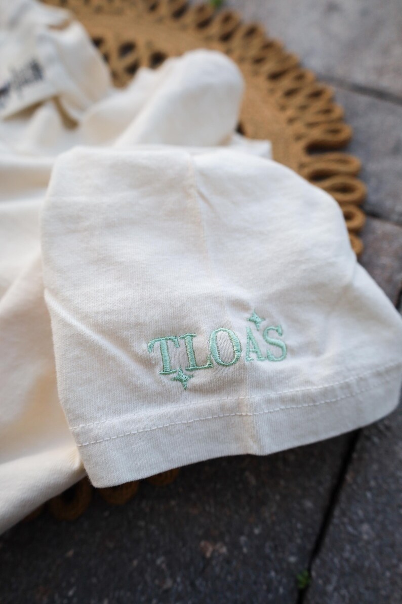 Puede incluir: Primer plano de una manga de camiseta color crema. El texto "TLOAS" est&aacute; bordado en color verde claro en la manga. La camiseta est&aacute; doblada y el fondo est&aacute; borroso.