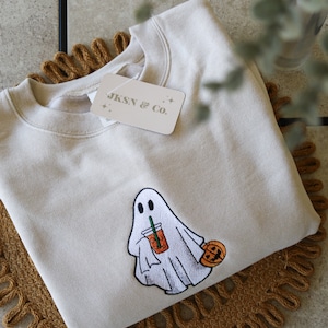 May include: A beige sweatshirt with an embroidered ghost holding a pumpkin spice latte and a small pumpkin. The sweatshirt has a tag that says "JKSN & Co."