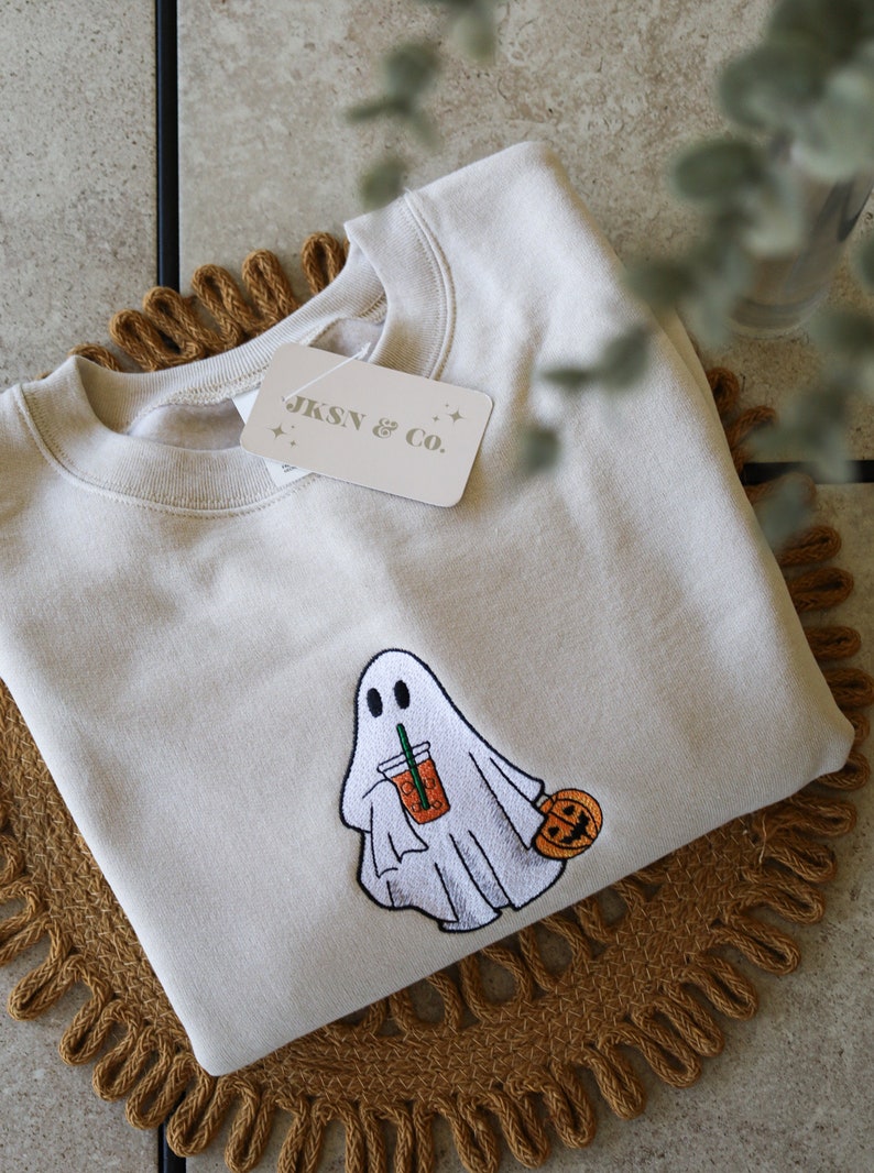 May include: A beige sweatshirt with an embroidered ghost holding a pumpkin spice latte and a small pumpkin. The sweatshirt has a tag that says "JKSN & Co."