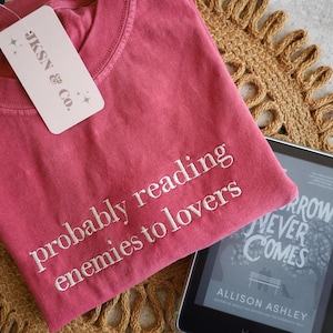 May include: A red t-shirt with the text "probably reading enemies to lovers" embroidered on the front. The t-shirt is lying on a woven mat with a Kindle e-reader on top of it. The Kindle is displaying the cover of the book "Tomorrow Never Comes" by Allison Ashley.