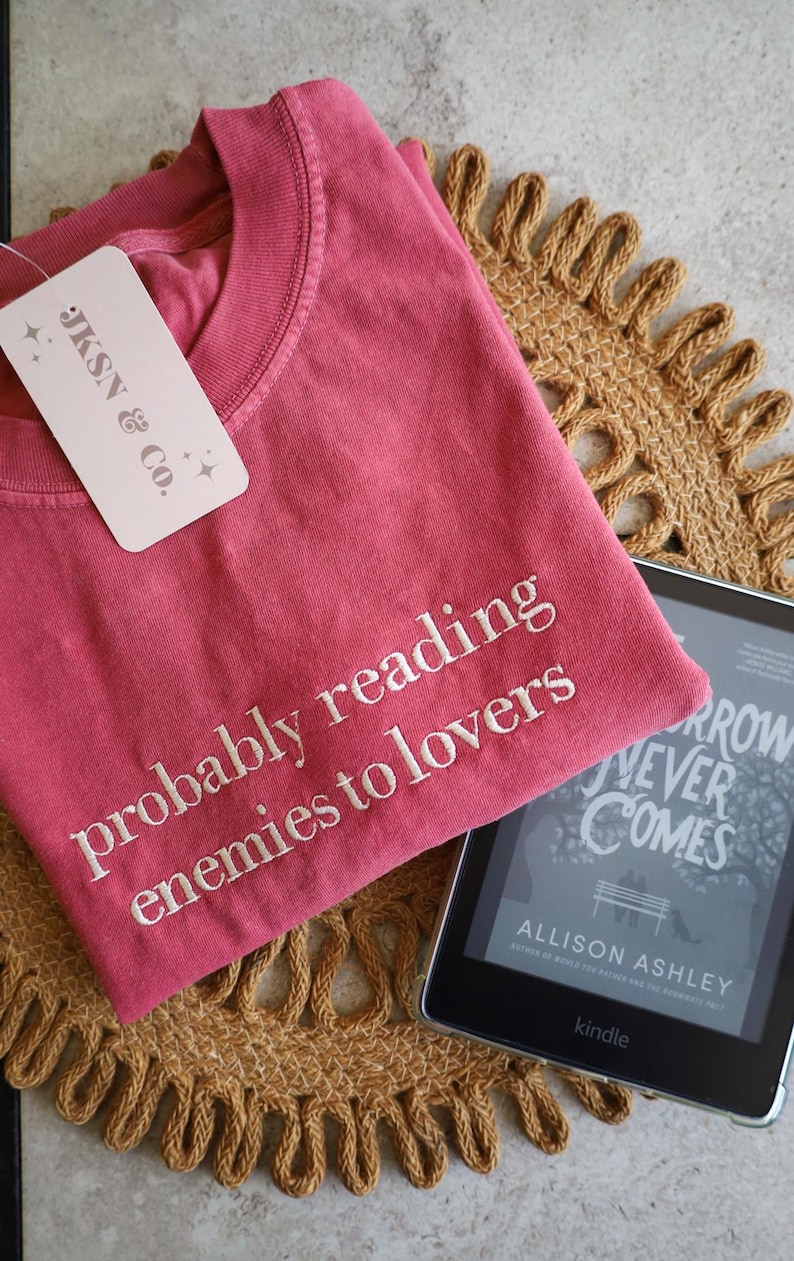 May include: A red t-shirt with the text "probably reading enemies to lovers" embroidered on the front. The t-shirt is lying on a woven mat with a Kindle e-reader on top of it. The Kindle is displaying the cover of the book "Tomorrow Never Comes" by Allison Ashley.