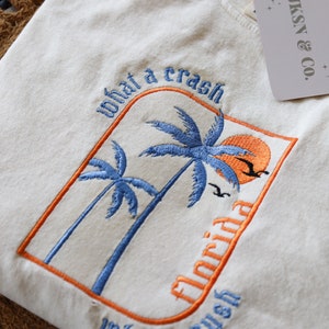 May include: A white t-shirt with an embroidered design of palm trees and a sunset. The text "what a crash" and "what a rush" is embroidered in blue and orange on the shirt. The text "Florida" is embroidered in blue on the shirt.