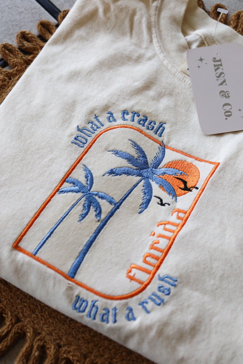May include: A white t-shirt with an embroidered design of palm trees and a sunset. The text "what a crash" and "what a rush" is embroidered in blue and orange on the shirt. The text "Florida" is embroidered in blue on the shirt.