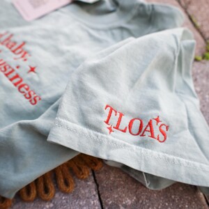 Puede incluir: Una camiseta verde claro con texto bordado en rojo que dice "baby + business" y "TLOA'S". La camiseta tiene una etiqueta adjunta y est&aacute; doblada, con el detalle de la manga visible. La camiseta est&aacute; sobre una superficie texturizada.