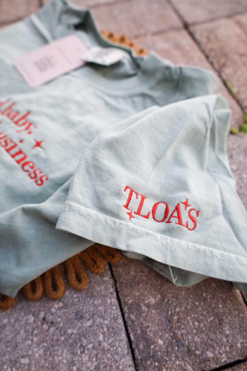 Puede incluir: Una camiseta verde claro con texto bordado en rojo que dice "baby + business" y "TLOA'S". La camiseta tiene una etiqueta adjunta y est&aacute; doblada, con el detalle de la manga visible. La camiseta est&aacute; sobre una superficie texturizada.