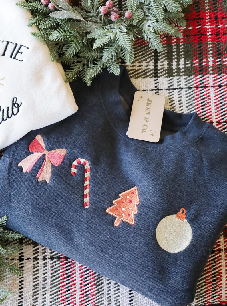 Może przedstawiać: Granatowa bluza z haftowanymi wzorami o tematyce świątecznej, w tym kokardą, laską cukrową, choinką i ozdobą. Bluza ma metkę z napisem "JKSN & Co."