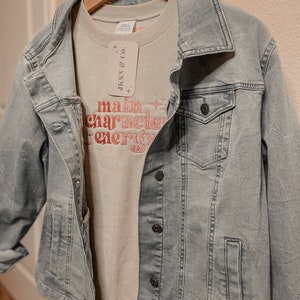 May include: A light gray denim jacket with a light gray sweatshirt underneath. The sweatshirt has the text "main character energy" in pink lettering. A white tag with the text "JKSNY & CO!" is attached to the sweatshirt.